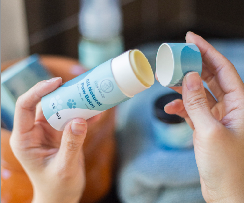 Two hands holding open tubes of All Natural Paw Balm with a blurred background.