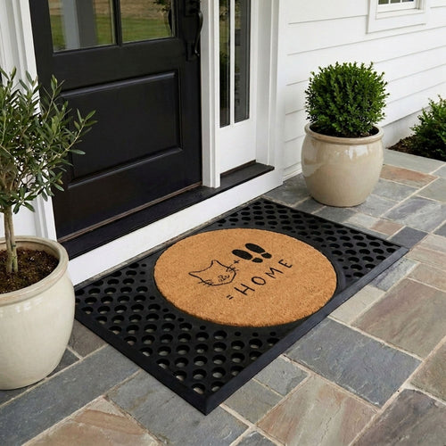 Doormat with Cat + Feet = Home doormat featuring a round natural coir insert with cat face and footprints design, surrounded by a black rectangular rubber frame with circular cutouts on a stone patio in front of a black door.