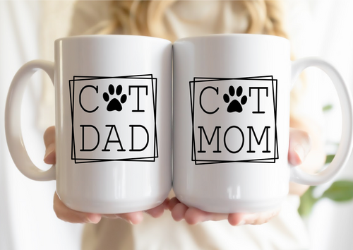 Two white mugs with 'Cat Dad' and 'Cat Mom' text held by a person.
