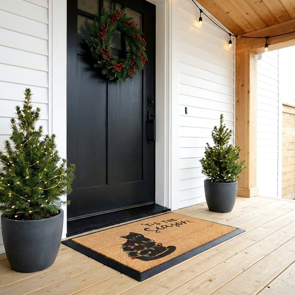 Front door of a house with a wreath, potted plants, and a doormat with a Christmas design.