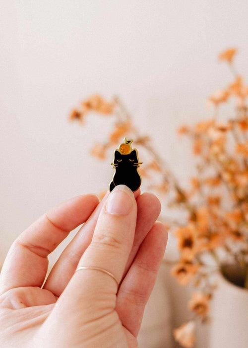 Hand holding a small black cat figurine with a gold collar against a blurred background of flowers.
