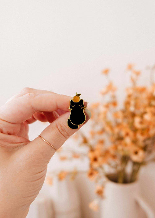 Hand holding a black cat-shaped earring with a blurred background of flowers and a vase.
